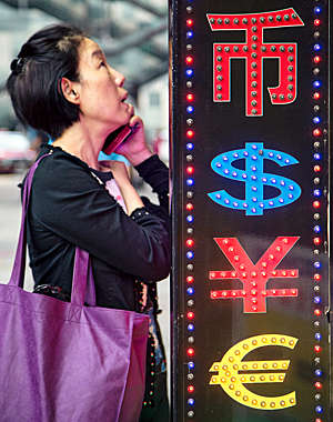 Asian woman talks on her smartphone next to a neon sign displaying currency codes.