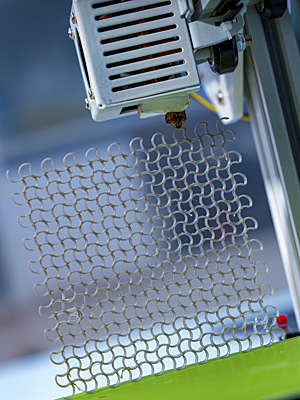 3D printer in the process of printing a square-shaped metal-looking grid.