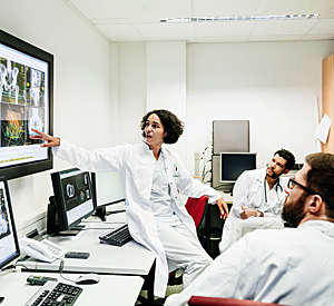 A clinical registrar reviewing a patients test results, looking at some information on a large monitor with some other doctors.