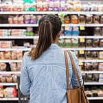 In this rear view, an unrecognizable woman stands with a shopping cart in front of a shelf full of food in the bread aisle of a grocery store.